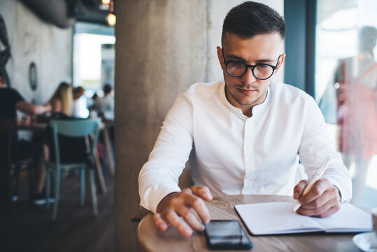 Serious man using smartphone and taking notes in cafe