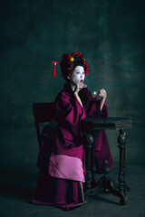 Tea drinking. Young japanese woman as geisha isolated on dark green background. Retro style, comparison of eras concept. Beautiful female model like bright historical character, old-fashioned.