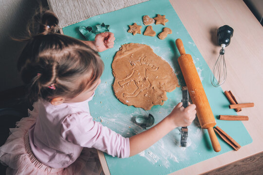 Cute adorable toddler girl cutting cookies from fresh dough  and baking gingerbread Christmas biscuits at the kitchen. Genuine authentic lifestyle moments. Real life. Winter holidays concept.