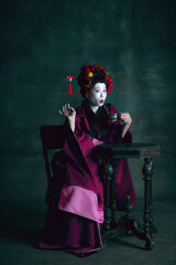 Having fun. Young japanese woman as geisha isolated on dark green background. Retro style, comparison of eras concept. Beautiful female model like bright historical character, old-fashioned.