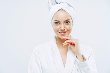 Studio shot of pretty beauty woman has washed hair, wears wrapped towel on head, has manicure, cute natural face, touches chin gently, looks with tender smile, dressed in bath robe, poses indoor