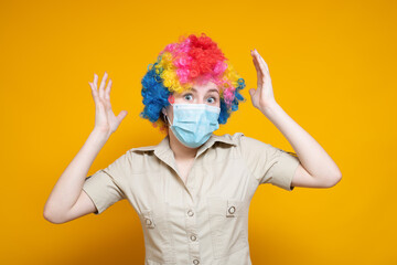 Emotional woman with rainbow wig and clown nose in medical mask on a yellow background. Holidays in a pandemic