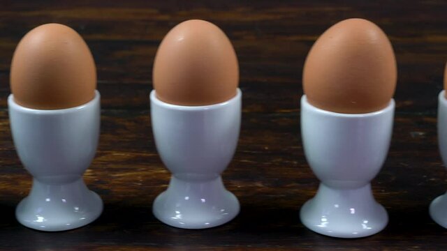 Panning shot across a wooden table to one boiled egg in a white egg cup that has a smiling face drawn on it
