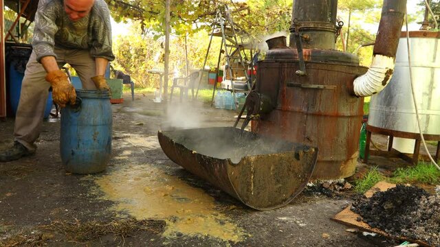 Man is transferring used fruit marc made of chopped apples, after distillation, using plastic bucket for filling small barrel to throw it as garbage. 