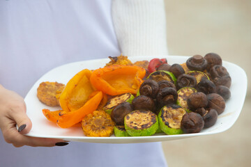 Hands hold a plate of grilled vegetables and mushrooms.