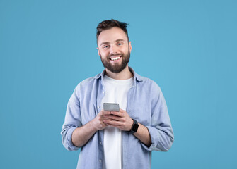 Portrait of cool bearded guy using mobile phone on blue studio background