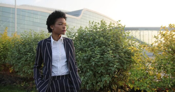 4k. Slow motion. Serious prety Afro-American businesswoman crosses her arms on chest standign before green bushes in the rays of evening sun