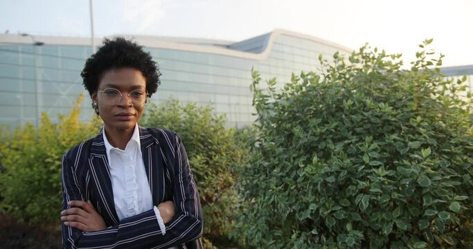 4k. Slow motion. Serious prety Afro-American businesswoman crosses her arms on chest standign before green bushes in the rays of evening sun