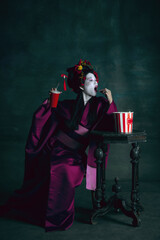 Fototapeta na wymiar Fastfood lover. Young japanese woman as geisha isolated on dark green background. Retro style, comparison of eras concept. Beautiful female model like bright historical character, old-fashioned.