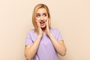 young blonde woman feeling happy, excited and surprised, looking to the side with both hands on face