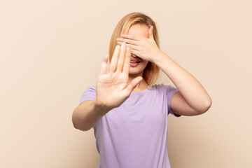 young blonde woman covering face with hand and putting other hand up front to stop camera, refusing...