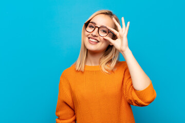 young blonde woman smiling happily with funny face, joking and looking through peephole, spying on...