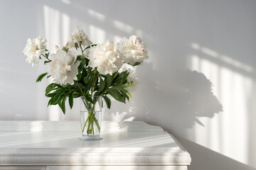 white peonies in a vase on a white vintage table and beautiful shadows on the wall - copy space © diyanadimitrova