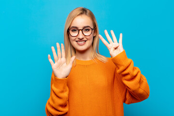 young blonde woman smiling and looking friendly, showing number nine or ninth with hand forward,...