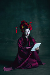 Scrolling tablet. Young japanese woman as geisha isolated on dark green background. Retro style, comparison of eras concept. Beautiful female model like bright historical character, old-fashioned.