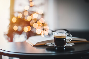 Open book, coffee and glasses on table. In the background a Christmas tree and Christmas lights. Christmas atmosphere.