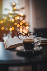 Open book, coffee and glasses on table. In the background a Christmas tree and Christmas lights. Christmas atmosphere.