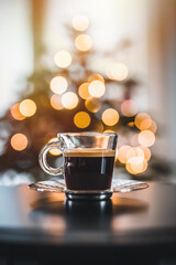 Obraz premium Glass cup of coffee close up. In the background a Christmas tree and Christmas lights.