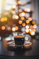 Glass cup of coffee close up. In the background a Christmas tree and Christmas lights.