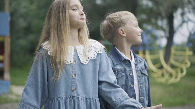 Excited girl talking and gesturing as shy boy kissing her on cheek. Romantic Caucasian children chatting outdoors on cloudy summer day. First love romance and dating concept.