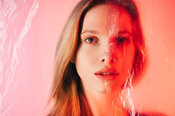 Skin rejuvenation. Aesthetic cosmetology. Lifting contouring. Art portrait of woman half face covered with wrinkled transparent polyethylene film in red neon light isolated on blur pink background.