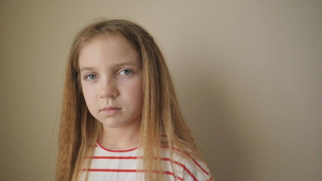 Little tired girl with long blonde hair posing to camera and swinging her body from side to side at room. Portrait of small bored child against the background of beige wall. Close up Slow motion