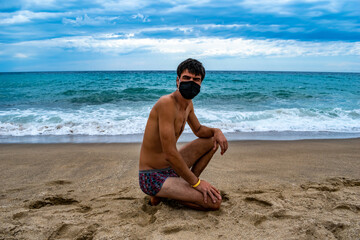 Fototapeta na wymiar Young dark-haired man in swimming trunks and a protective medical face mask sits on the sand against the backdrop of turquoise sea water. Concept of a safe tourist holiday during covid-19 quarantine