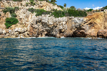 Fototapeta na wymiar Rocky sandstone coast in Alanya (Turkey). Beautiful seascape with blue-turquoise water, brown-gray-yellow cliff and green plants growing on the bluff slopes