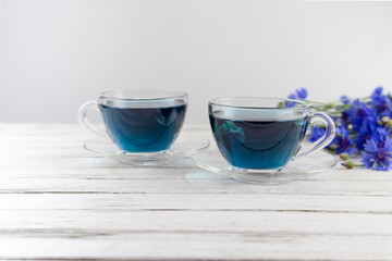glass cups and teapot of Butterfly pea tea. Anchan, clitoria ternatea on a white wood background. with bouquet of blue flowers.