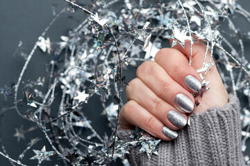 Female hand in gray knitted sweater with beautiful holiday manicure - silver glittered nails with Christmas twisted wire with stars. Selective focus. Closeup view. Blurred background with copy space