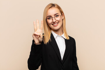 young blonde woman smiling and looking friendly, showing number three or third with hand forward,...