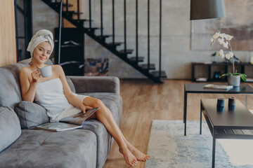 Happy young woman poses wrapped in bath towel, drinks aromatic coffee and reads magazine, sits on comfortable sofa, poses in living room at home. Domestic atmoshpere, leisure time, lifestyle