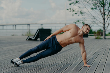 Fototapeta na wymiar Sporty bodybuilder stands in side plank, exercises outdoor and wears sport clothes, leads healthy lifestyle, has fit body and muscles. Outdoor shot of determined sportsman does health activities