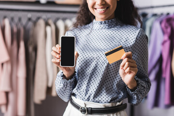 cropped view of african american proprietor holding credit card and smartphone with blank screen in clothing shop