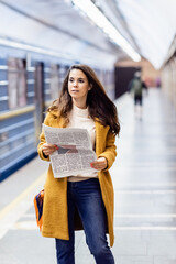 young woman in autumn outfit holding newspaper near blurred metro train on platform