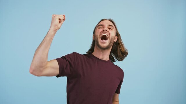 Male rubbing folded hands, screaming yes raising clenched fists up being overjoyed, posing on blue background
