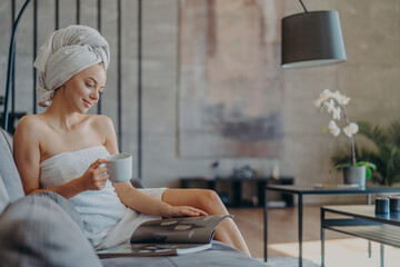 Beautiful relaxed European woman with natural beauty wrapped in towel, sits on comfortable sofa, drinks tea and reads magazine, enjoys spending time at home. Women, comfort, hygiene day off concept