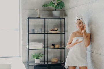 Beautiful European woman applies face mask, has flawless skin and looks somewhere thoughtfully, uses beauty cosmetics, stands wrapped in towel at cozy bathroom. Young lady applies body cream