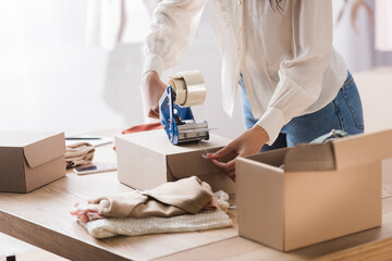 Cropped view of african american businesswoman packing box with adhesive tape near clothes on blurred foreground in showroom