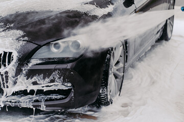 Washing auto at manual car wash. Black vehicle covered with water and washing foam cleaned by high pressured water stream
