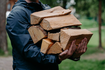 Bijgesneden schot van anonieme man in zwarte jas draagt stapel brandhout poses tegen wazig bos achtergrond.
