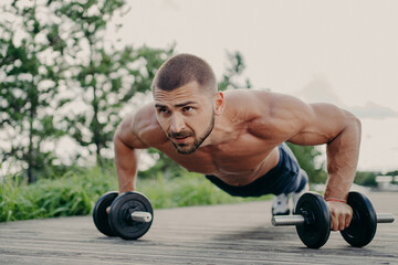 Young motivated sportsman stands in plank pose, trains muscles with barbells, focused into distance...