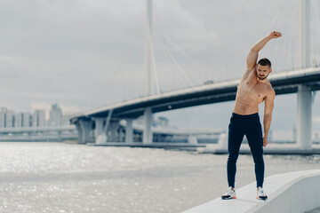 Fototapeta na wymiar Full length shot of muscles active man does sport exercises outdoor poses near river bridge wears trousers and sneakers has beautiful male torso achieves best shape. Healthy lifestyle concept