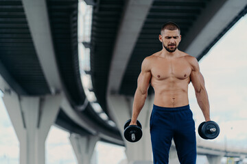 Fototapeta na wymiar Serious bearded European man with sporty body prepares for barbell workout stands under bridge outdoor has strong biceps and arms concentrated down. Bodybuilding power lifting training concept