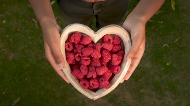 Young woman lowering a heart shaped wooden bowl of fresh red raspberries to camera