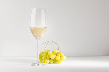 White wine in a glass goblet and a bunch of grapes on a white table.