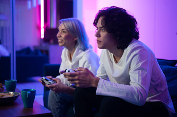 Couple of young adults playing video games at home. Emotional diverse gamers holding joysticks and compete in intense video game on gaming console.
