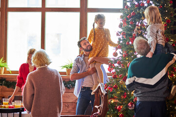 father and child girl decorating xmas tree while other family members decorating dinner table, on the eve of New Year, they smile, enjoy and relax after hard year