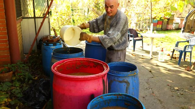 Farmer is transfuse fruit marc with plastic bucket.
Man is transferring fruit marc made of chopped apples using plastic bucket form one barrel to another. 
