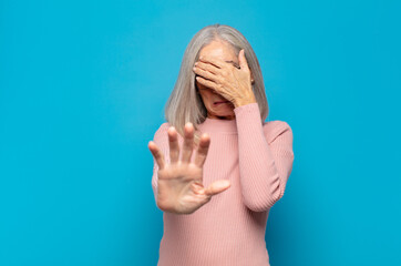 middle age woman covering face with hand and putting other hand up front to stop camera, refusing...